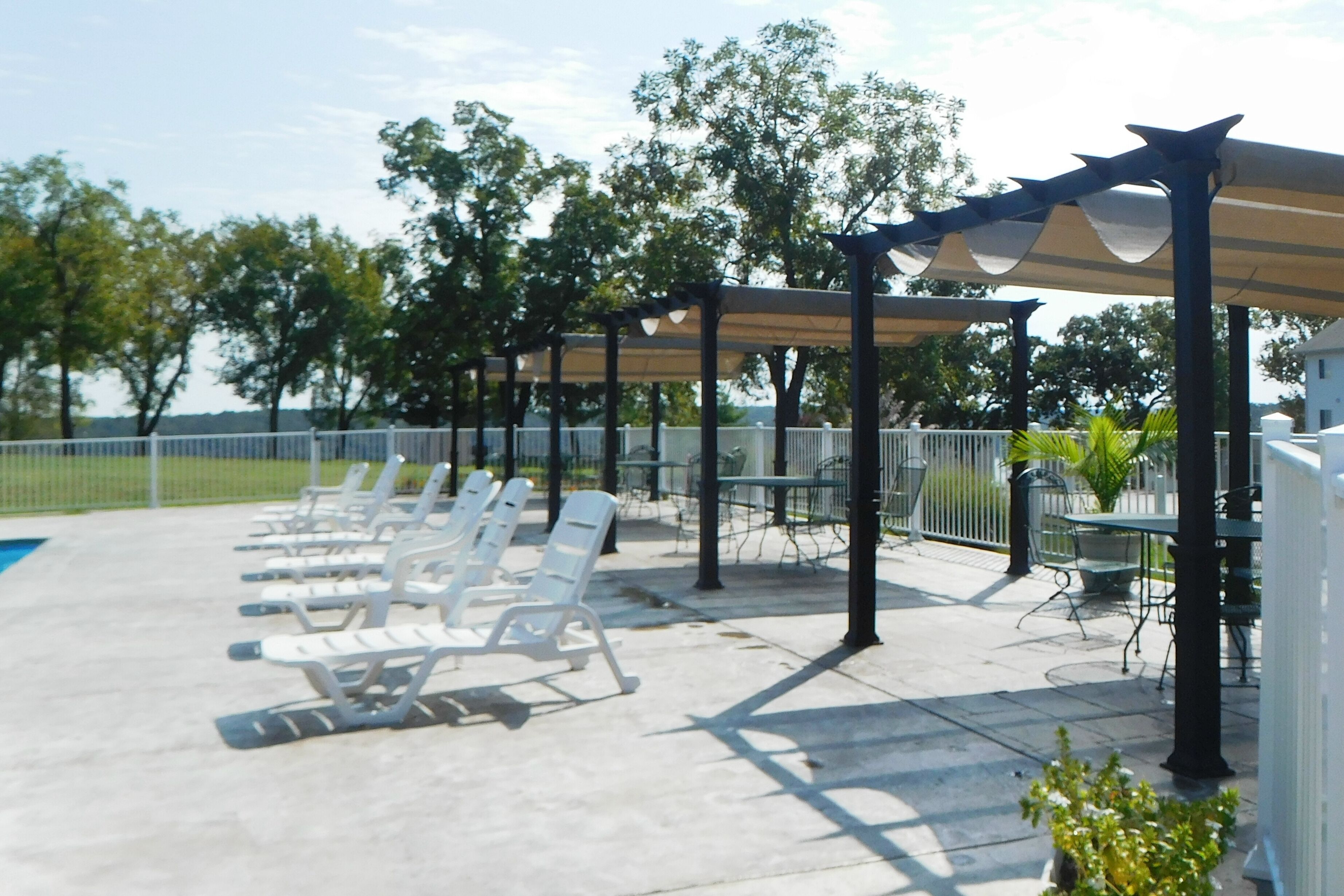 Un espace extérieur aménagé, abrité et doté de chaises longues et de tables blanches, entouré d’une clôture et d’arbres.