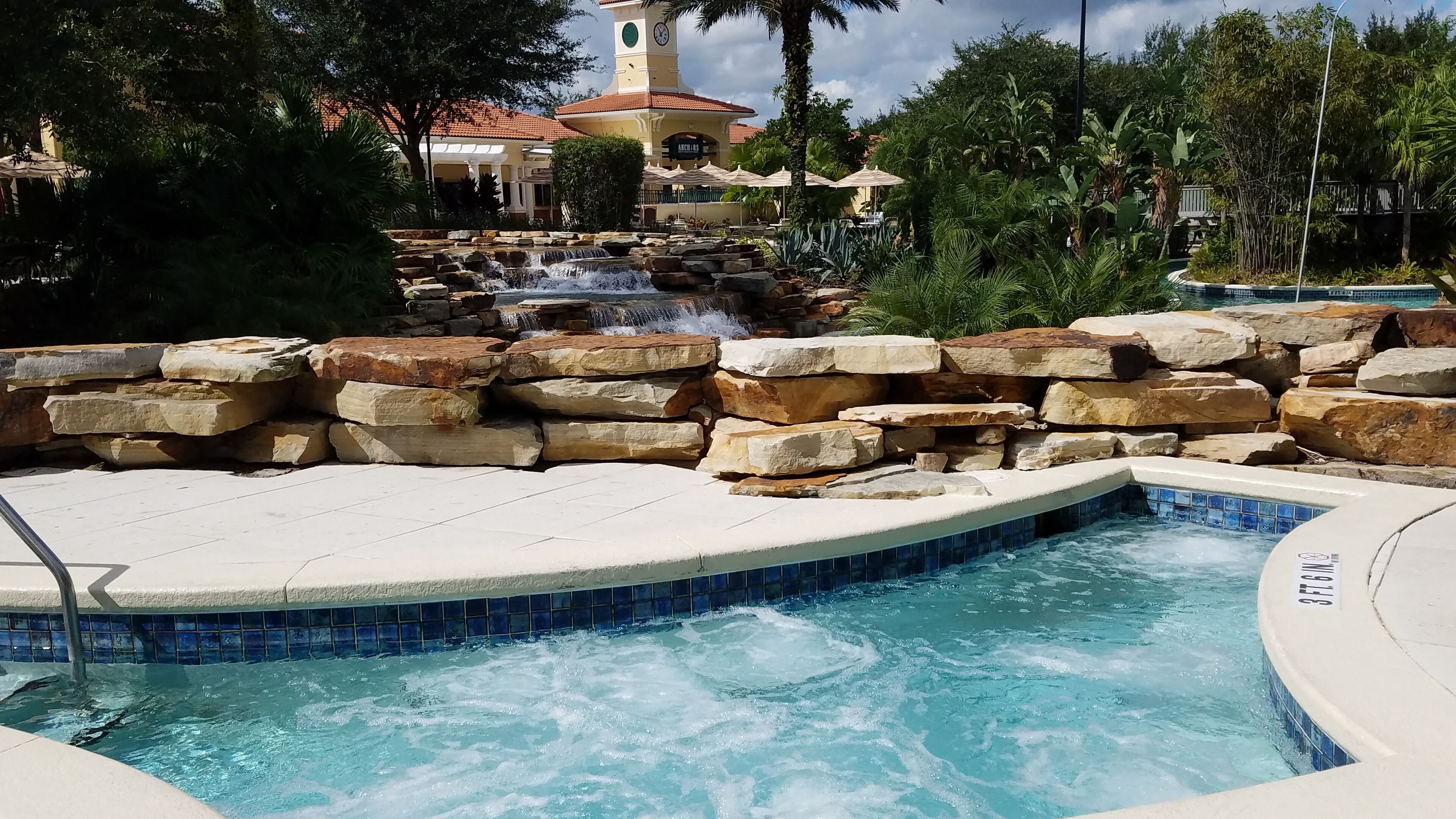 A swimming pool with a waterfall feature, surrounded by stone walls and tropical plants.