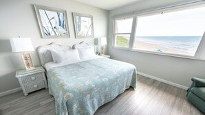 Une chambre avec un grand lit, une table de chevet, une commode et une fenêtre donnant sur la plage.
