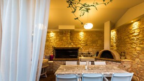 A stone-walled kitchen with a pizza oven, a dining table with chairs, and a hanging light fixture. - Dimitrios Villa, a Luxury Oasis with Pool & Gym (Rethymno)
