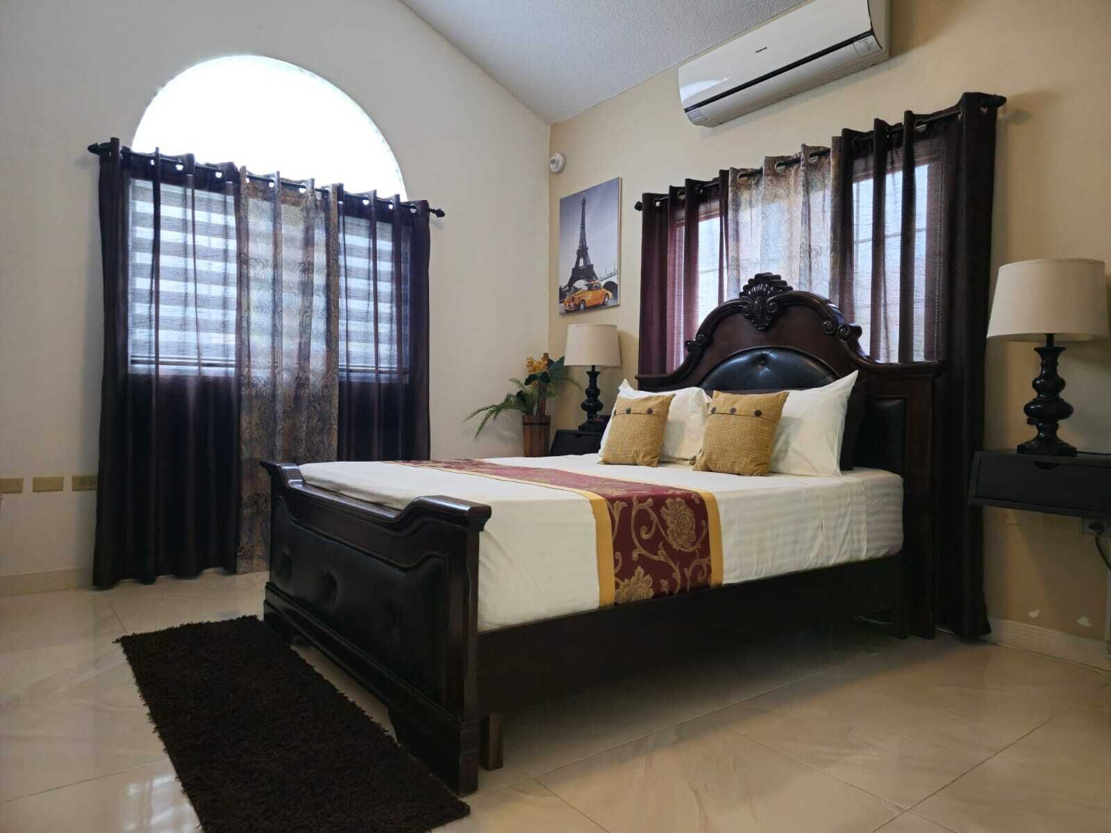 A bedroom with a large bed, dark curtains, a wooden headboard, a nightstand with a lamp, and a wall-mounted air conditioner.