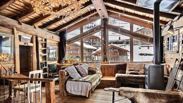 Interior rústico de una cabaña con paredes de madera, una zona de estar acogedora, chimenea y vistas a un pueblo nevado.