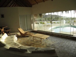 Una habitación con un sofá blanco curvo, una silla de mimbre y una mesa de madera. Un ventanal ofrece vistas a una piscina y zona de estar al aire libre.