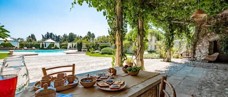 Uma área de refeições ao ar livre com uma mesa posta para uma refeição, cercada por vegetação e uma piscina ao fundo.