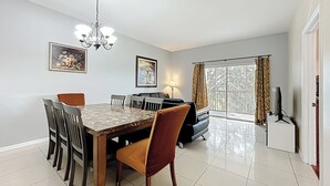 A dining area with a marble table, chairs, and a chandelier. There is a sofa and a TV in the background.
