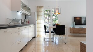 Una cocina moderna con armarios blancos, un comedor con mesa y sillas, un televisor sobre un mueble de madera y una ventana con vista a árboles.
