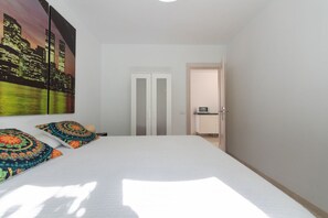 Un dormitorio con cama, dos almohadas, una mesita de noche, una puerta y vistas a un paisaje urbano.