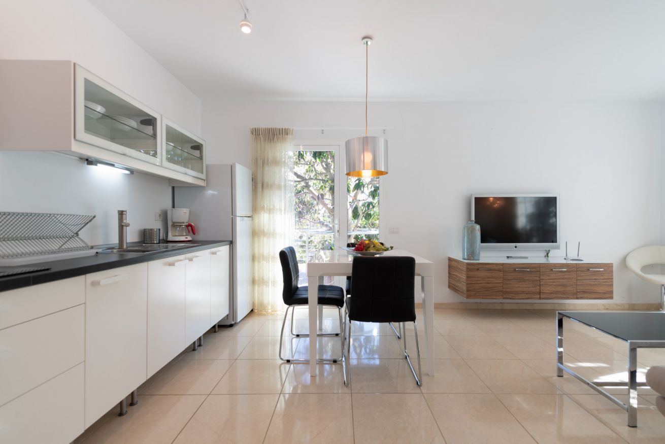 Une cuisine moderne avec des meubles blancs, un coin repas avec deux chaises, une table avec des fruits, un téléviseur posé sur un meuble en bois et une suspension décorative.