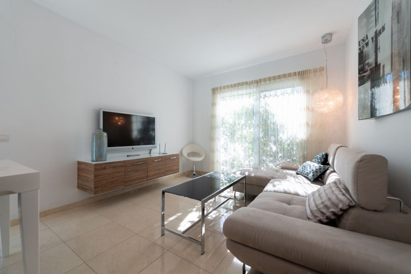 A modern living room with a sofa, a coffee table, a TV on a wooden cabinet, and a large window with curtains.