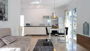 Una sala moderna con un sofá, una mesa de centro y un comedor con una mesa y sillas. Hay una zona de cocina con armarios y una ventana.