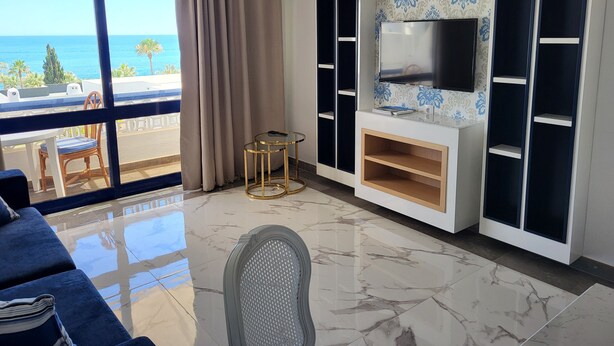 Un soggiorno moderno con una grande TV, un balcone con vista mare e un'area pranzo con tavolo e sedie.