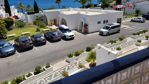 Un'area di parcheggio con diverse auto, un edificio bianco con archi e vista sul mare.