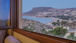 Une vue depuis une fenêtre donnant sur un paysage côtier, avec une colline, des bâtiments et une étendue d’eau.
