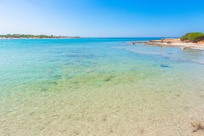 A beach with clear turquoise water, a sandy shore, and a distant shoreline with buildings.