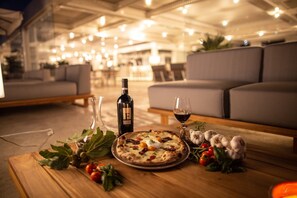 A dining table with a pizza, wine bottle, and glass, surrounded by fresh herbs and garlic cloves.