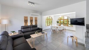 A modern living room with a black leather sofa, a wooden coffee table, and a TV mounted on the wall. There is a dining area with a white table and chairs, and large windows providing natural light. - This amazing 3 Bedroom villa is located in the center of protaras (Protaras)