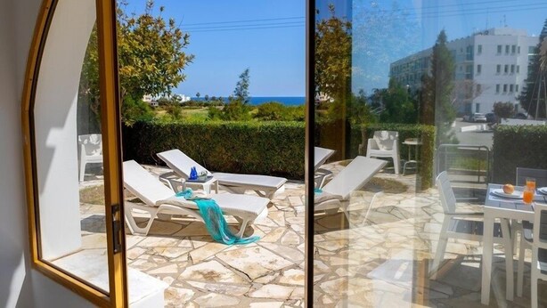 A sunlit patio with lounge chairs and a table, overlooking a landscaped garden and the sea. - This amazing 3 Bedroom villa is located in the center of protaras (Protaras)