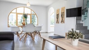 A modern living room with a sofa, a dining area with chairs and a table, a TV mounted on the wall, and a staircase. - This amazing 3 Bedroom villa is located in the center of protaras (Protaras)