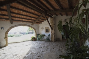 An outdoor patio area with stone flooring, a wooden ceiling, and a large archway leading to a garden. There are potted plants and a table with chairs. - Rustic farmhouse with sea views, large garden and private pool. (Platja d'Aro)
