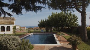 Piscina rodeada por um jardim com pérgola, um pequeno edifício com arcos e vista para o mar.