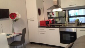 A compact kitchen with white cabinets, a microwave, a stove, and a sink. There's a small dining area with a chair and a table. -  BEAUTIFUL 2-ROOM APARTMENT "DESIGN" calm GROUND FLOOR LE LOGIS LE COL D'ASPIN (Lourdes)