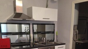 A compact kitchen with a stove, sink, and white cabinets. There is a dining table with a floral placemat and a red folder. -  BEAUTIFUL 2-ROOM APARTMENT "DESIGN" calm GROUND FLOOR LE LOGIS LE COL D'ASPIN (Lourdes)