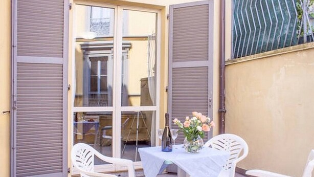Une terrasse extérieure avec une table dressée pour deux personnes, des chaises en plastique blanches et un vase rempli de fleurs.