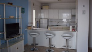 A kitchen with white cabinets, a refrigerator, a sink, and a TV on a shelf. There are three bar stools and a basket on the counter. - Front line apartment in Puerto Santiago-Los Gigantes with beautiful views.<br> (Puerto Santiago)