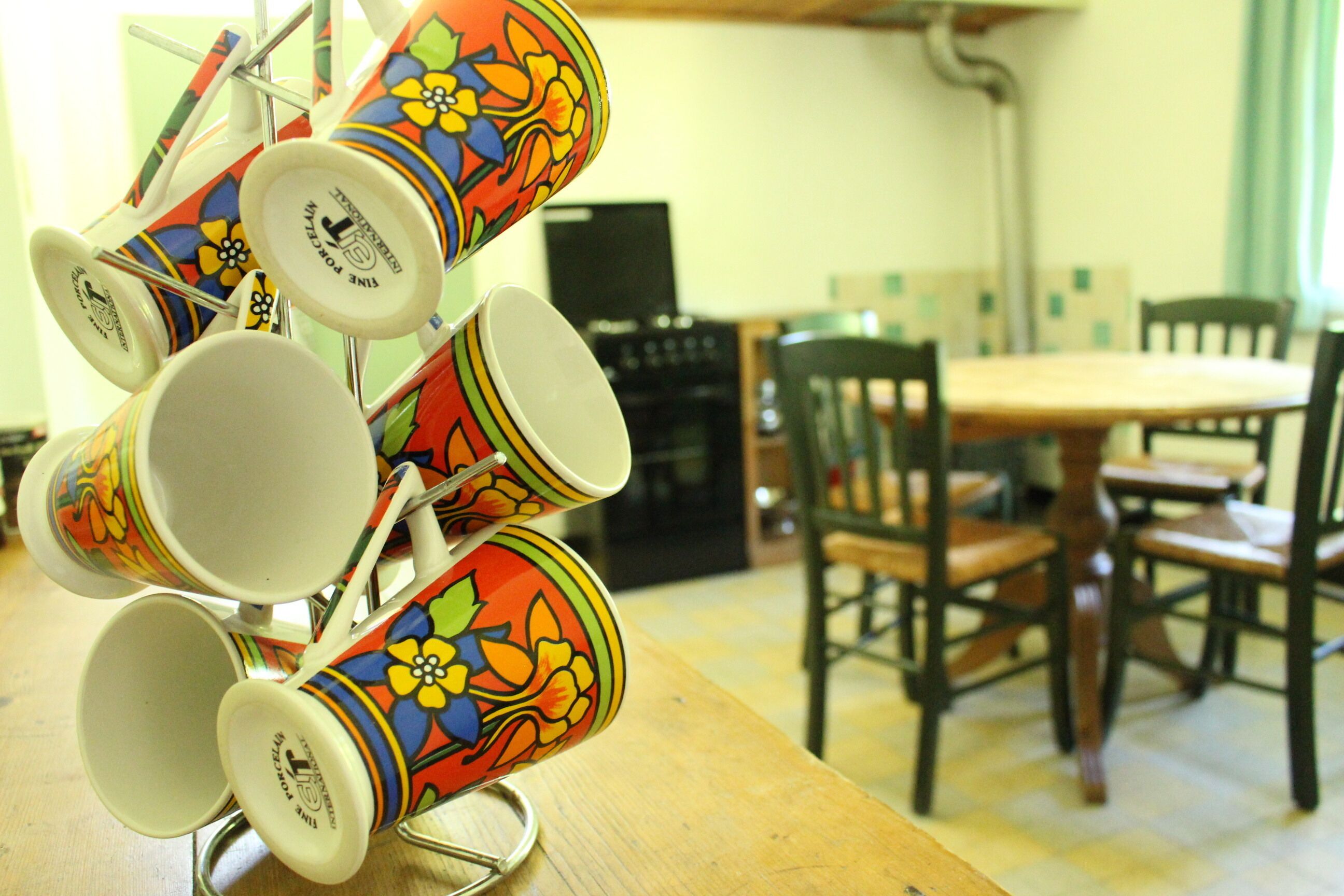 A dining area with a wooden table and chairs, a coffee press with colorful cups, and a television in the background.