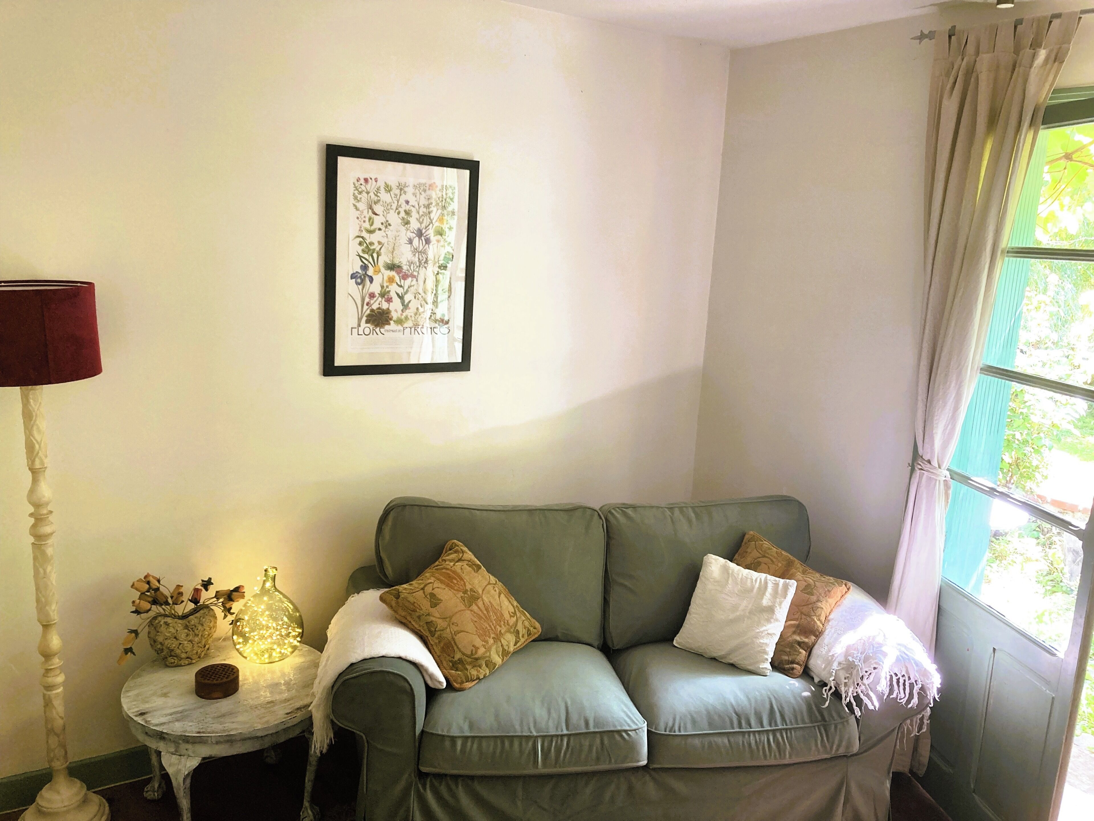 A living room with a green sofa, a small round table with a vase and a lamp, and a framed botanical print on the wall.