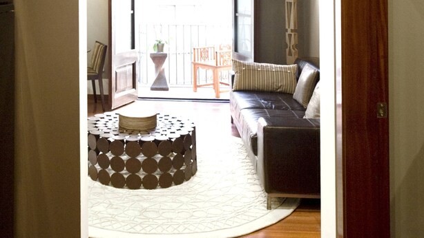 An interior with a sofa, a patterned ottoman, and a view through an arched doorway to an outdoor balcony.