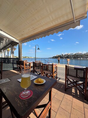 Um restaurante à beira-mar com mesas ao ar livre, vista para o mar e uma mesa com um copo de cerveja e batatas fritas.