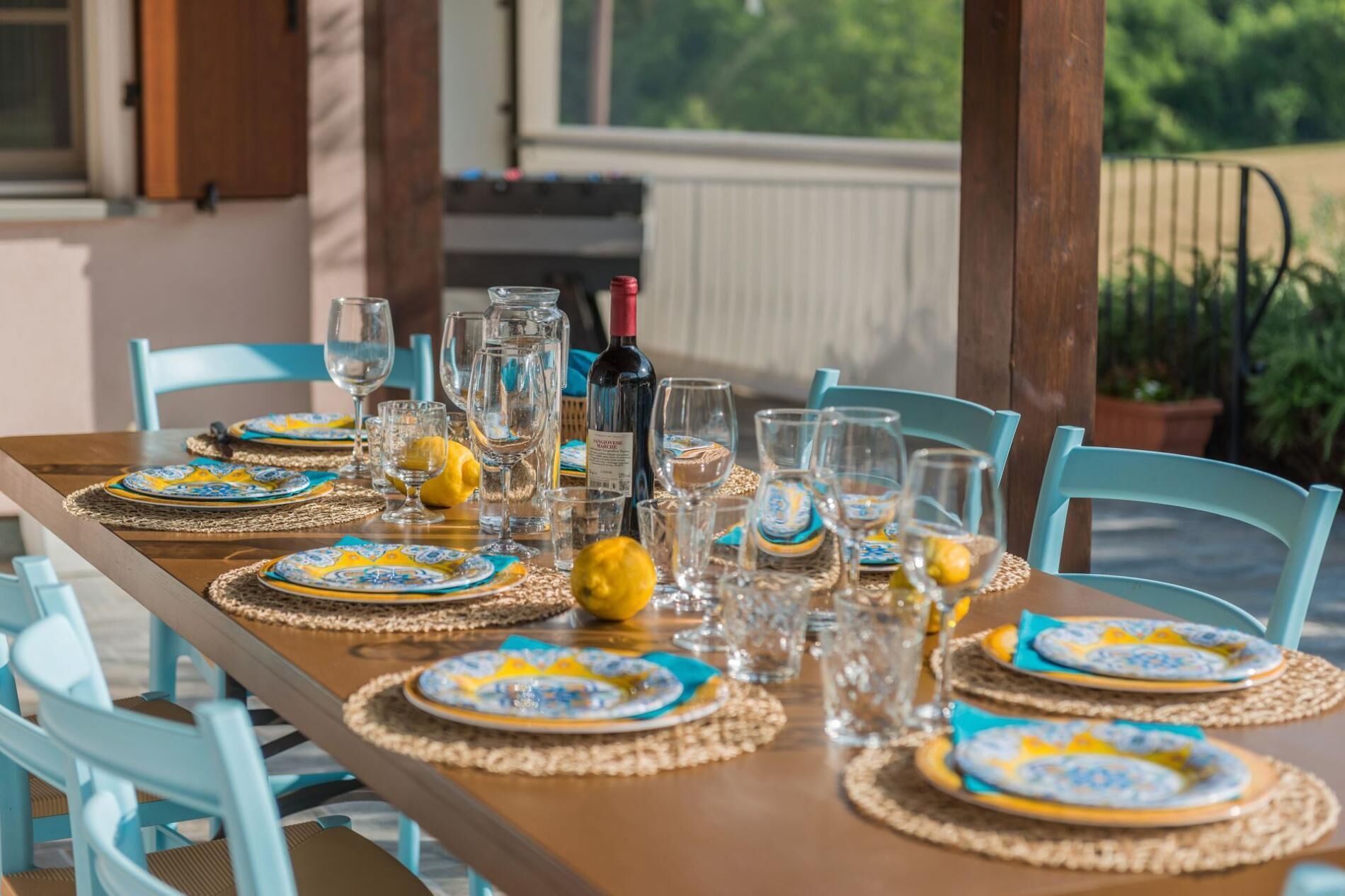 Een gedekte eettafel met borden, glazen en een fles, omringd door stoelen en een uitzicht op groen.