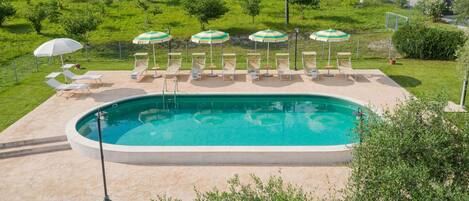 Piscina con sillas de descanso y sombrillas, rodeada de césped y árboles.
