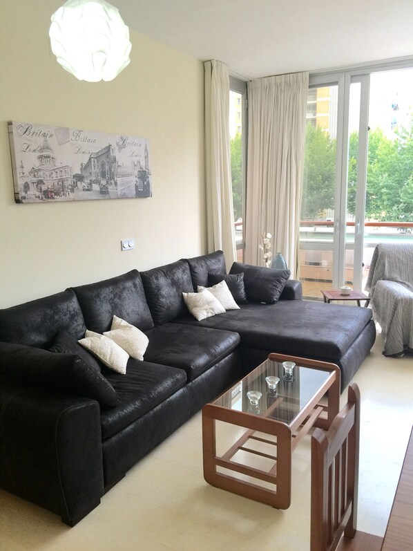 A modern living room with a black leather sofa, a glass coffee table, and a large window with curtains.