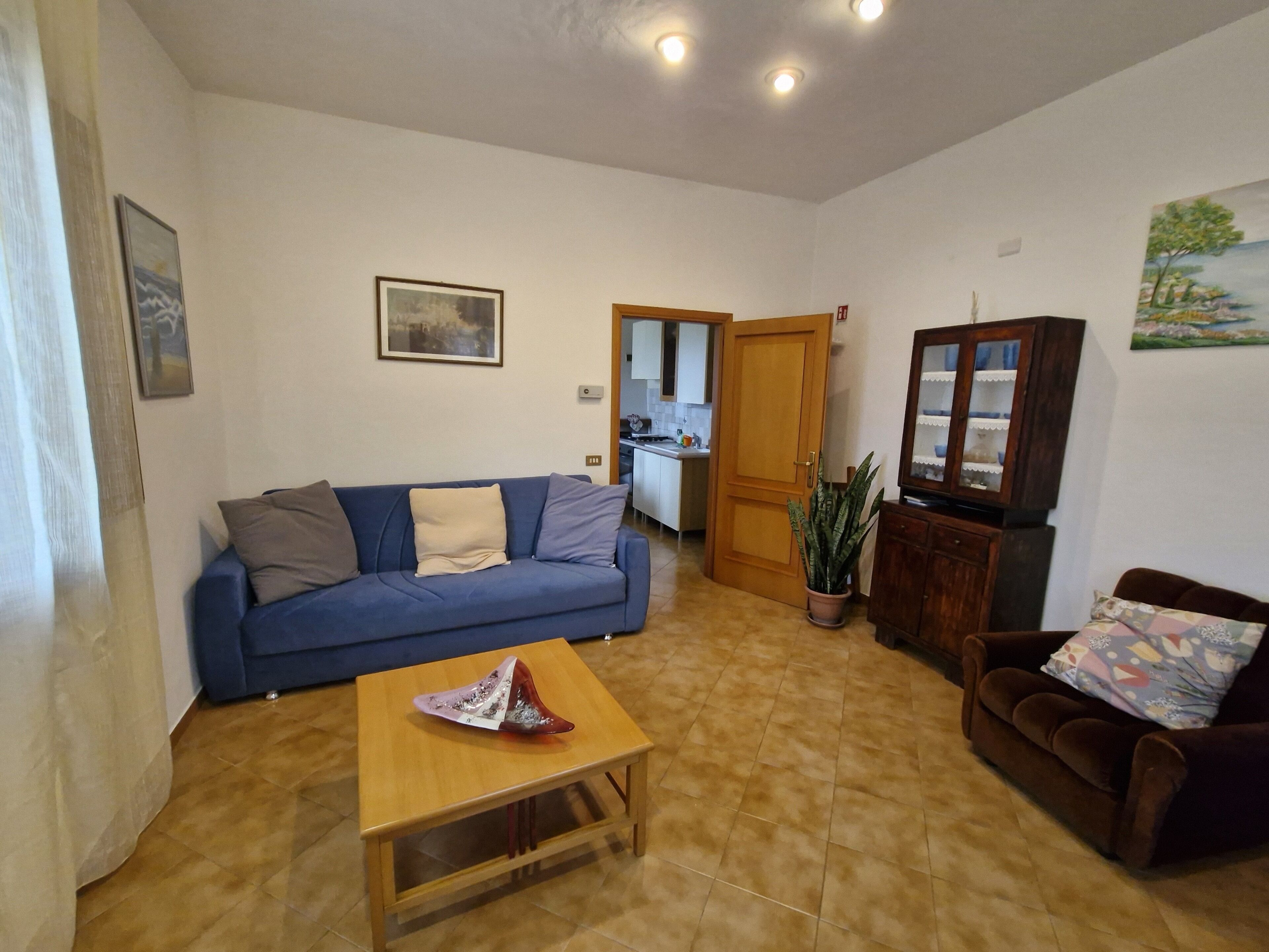 A living room with a blue sofa, a wooden coffee table, and a brown armchair. There is a kitchen visible through an open door in the background.