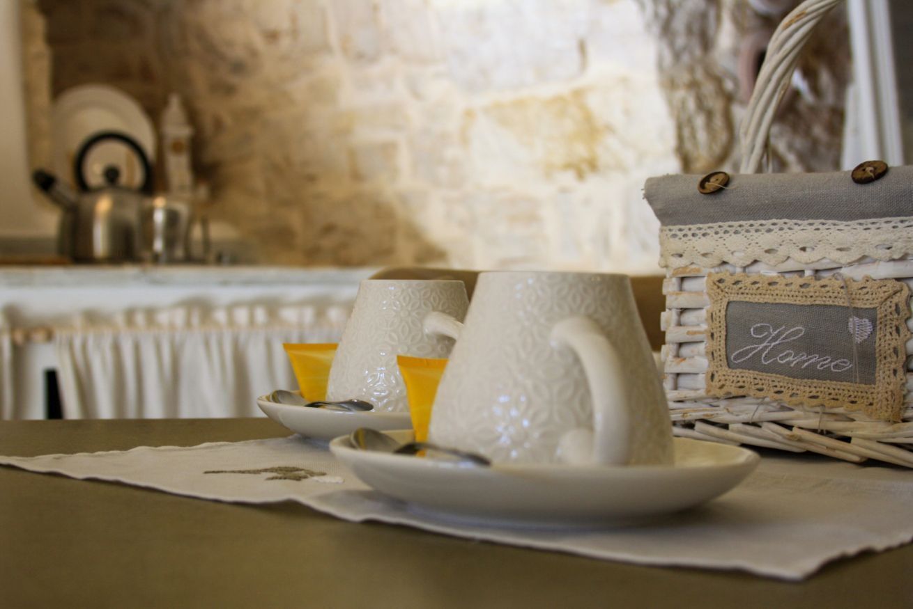 A table with white ceramic cups and saucers, a wicker basket with a label, and a rustic stone wall in the background.