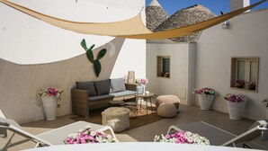 A sunlit patio with a sofa, ottoman, and lounge chairs, surrounded by potted plants and a large cactus.