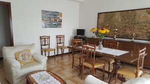 Zona pranzo con tavolo in legno, sedie e un vaso con girasoli. Si vede anche una televisione e un quadro di grandi dimensioni appeso al muro.