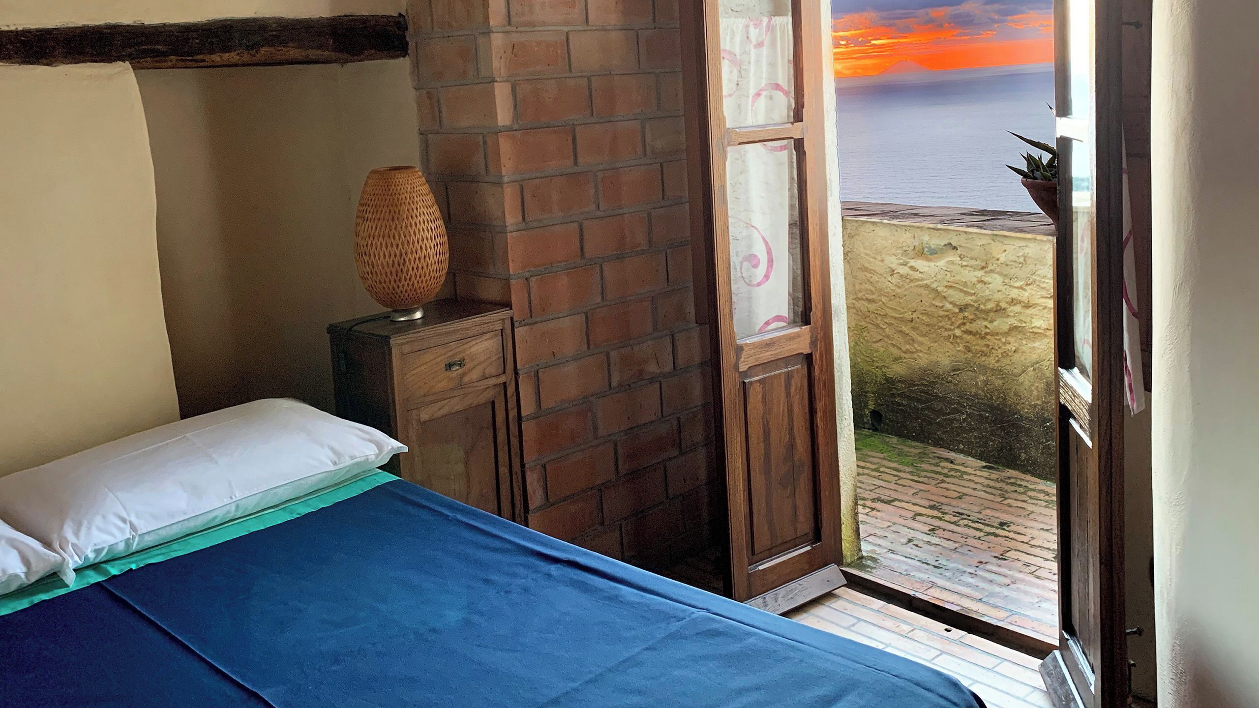 A bedroom with a bed, a nightstand, a lamp, and a view of the sea through an open door.