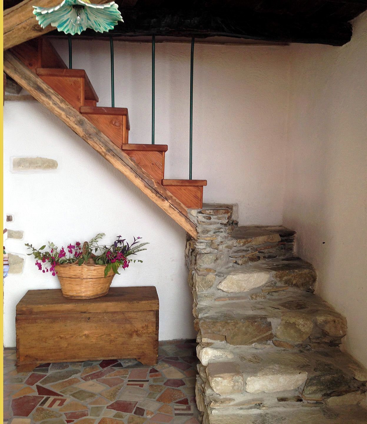 Eine Holztreppe mit einer Steinmauer auf der einen Seite und einer weißen Wand auf der anderen. Am Fuß der Treppe befindet sich eine Holzbank mit einem Blumenkorb.