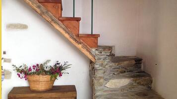 Eine Holztreppe mit einer Steinmauer auf der einen Seite und einer weißen Wand auf der anderen. Am Fuß der Treppe befindet sich eine Holzbank mit einem Blumenkorb.