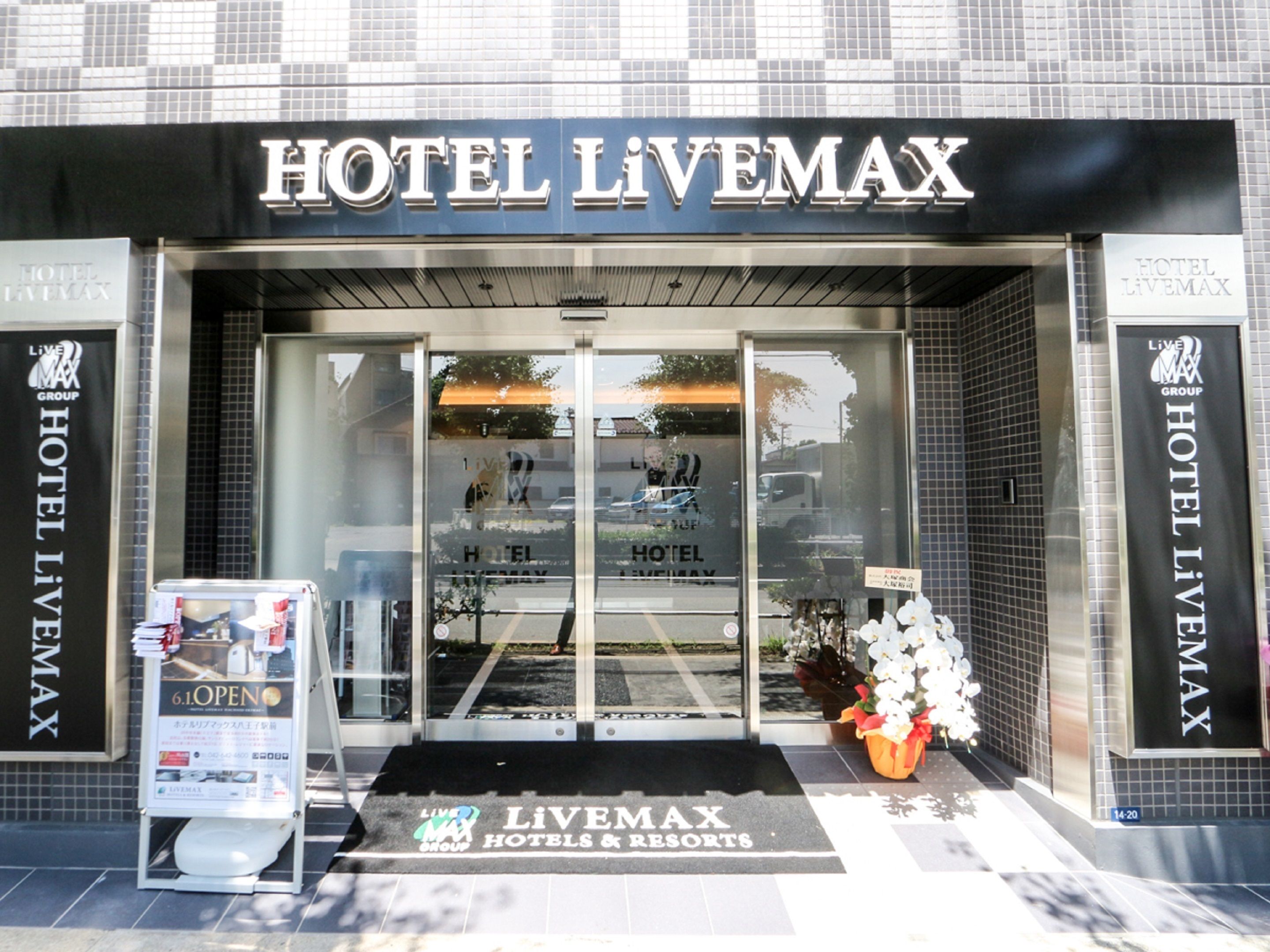 The entrance to Hotel Livemax with a clear glass facade, a black awning, and a welcome mat at the door.