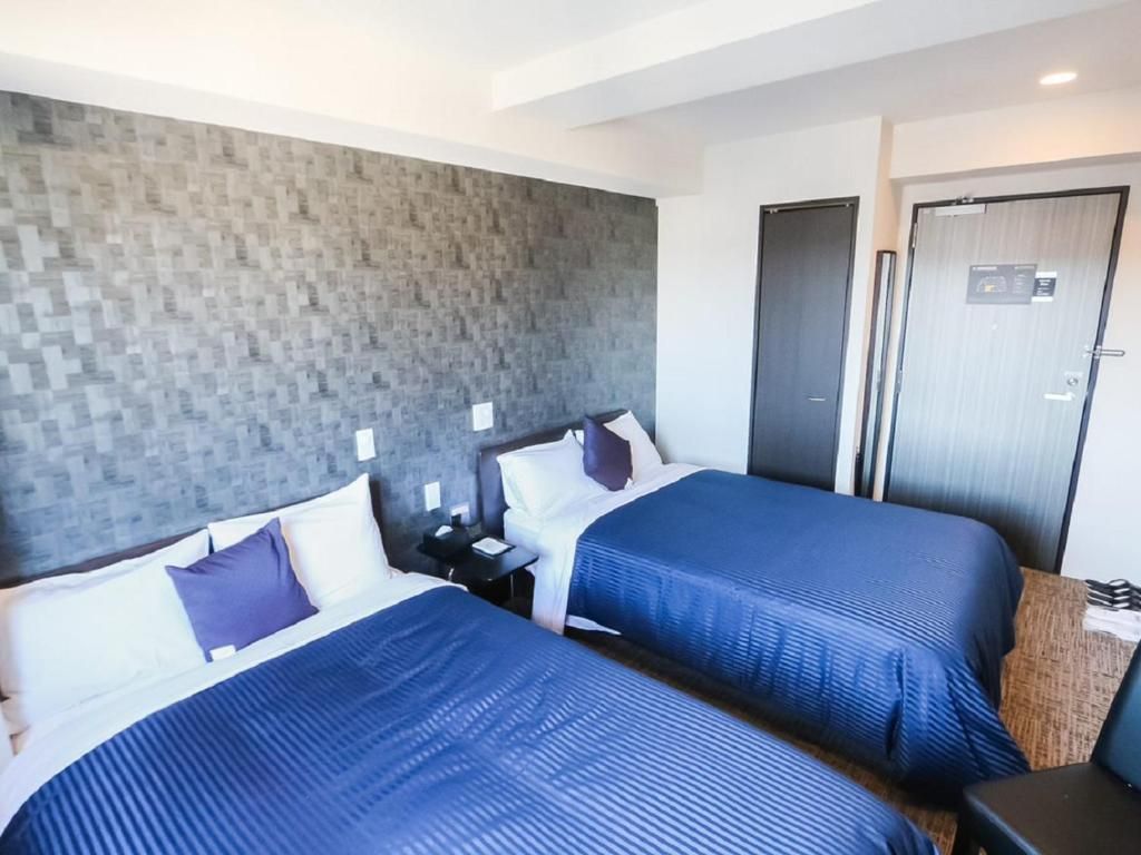 A hotel room with two beds, a grey textured headboard, a wooden nightstand, and a door with a digital display.
