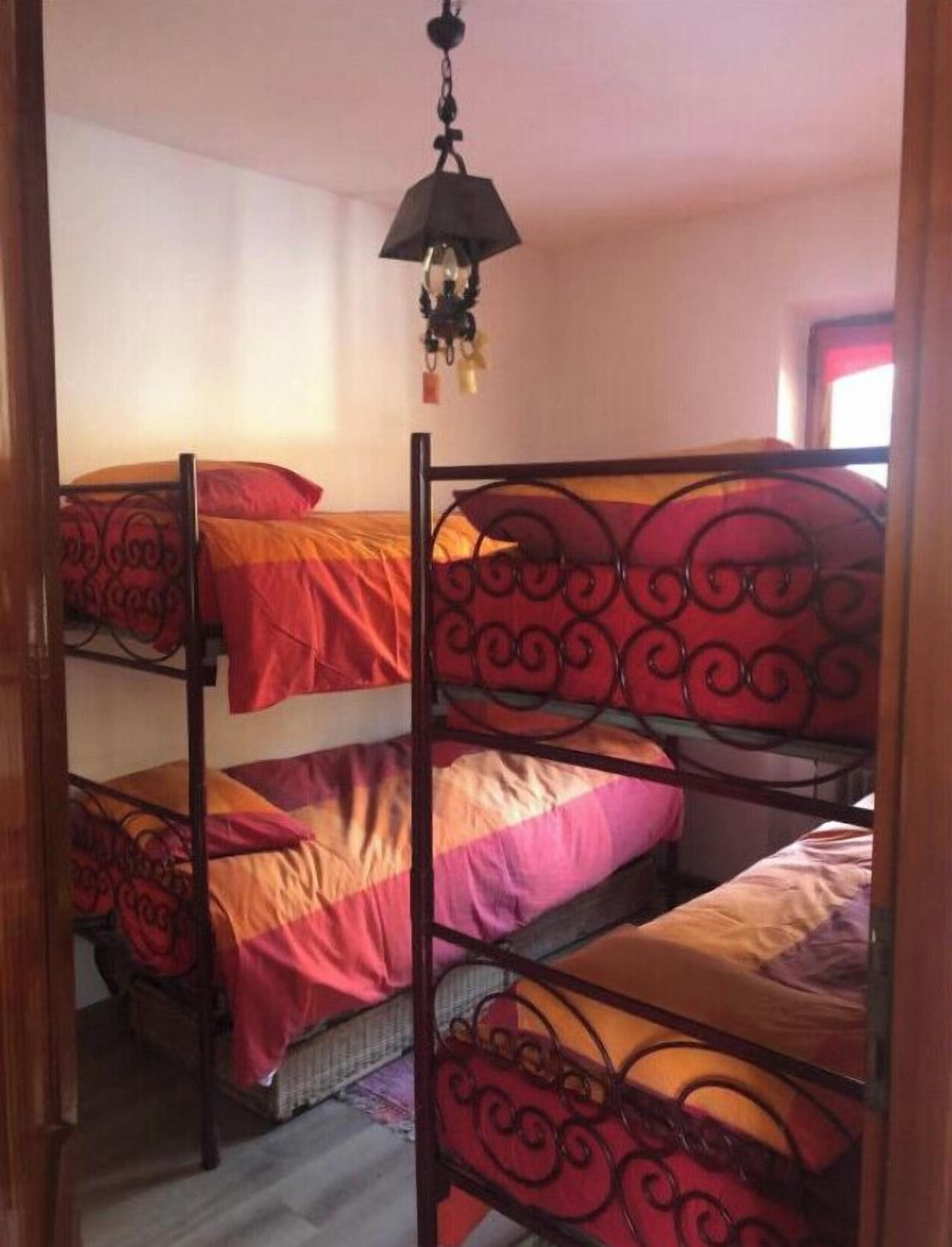 Una habitación con tres literas, cada una con una colcha roja y naranja. Hay una lámpara colgante sobre las camas.