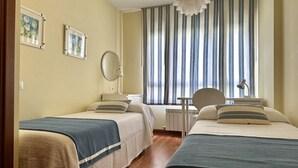 A hotel room with two beds, a bedside table with a lamp, a radiator, and a window with striped curtains.