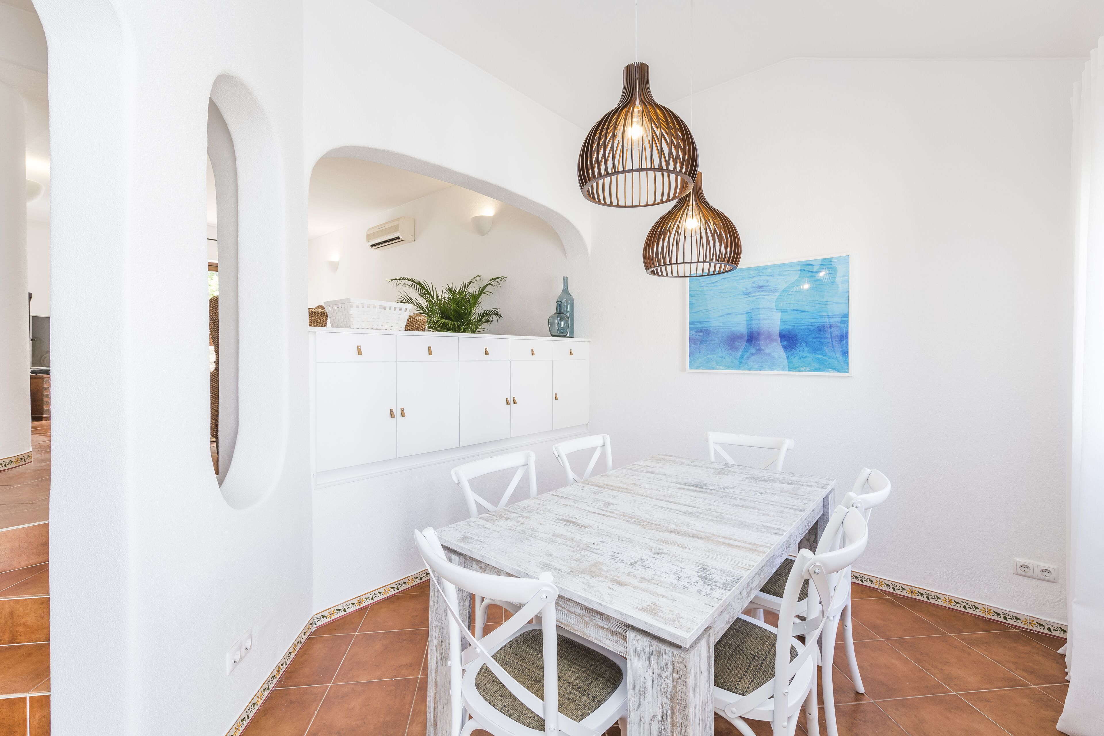 Un coin repas avec une table et des chaises blanches, un meuble blanc orné d’une plante, et un tableau abstrait bleu.