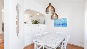 A dining area with a white table and chairs, a white cabinet with a plant, and a blue abstract painting. - CASAS DA PRAIA,   55 (Almancil)