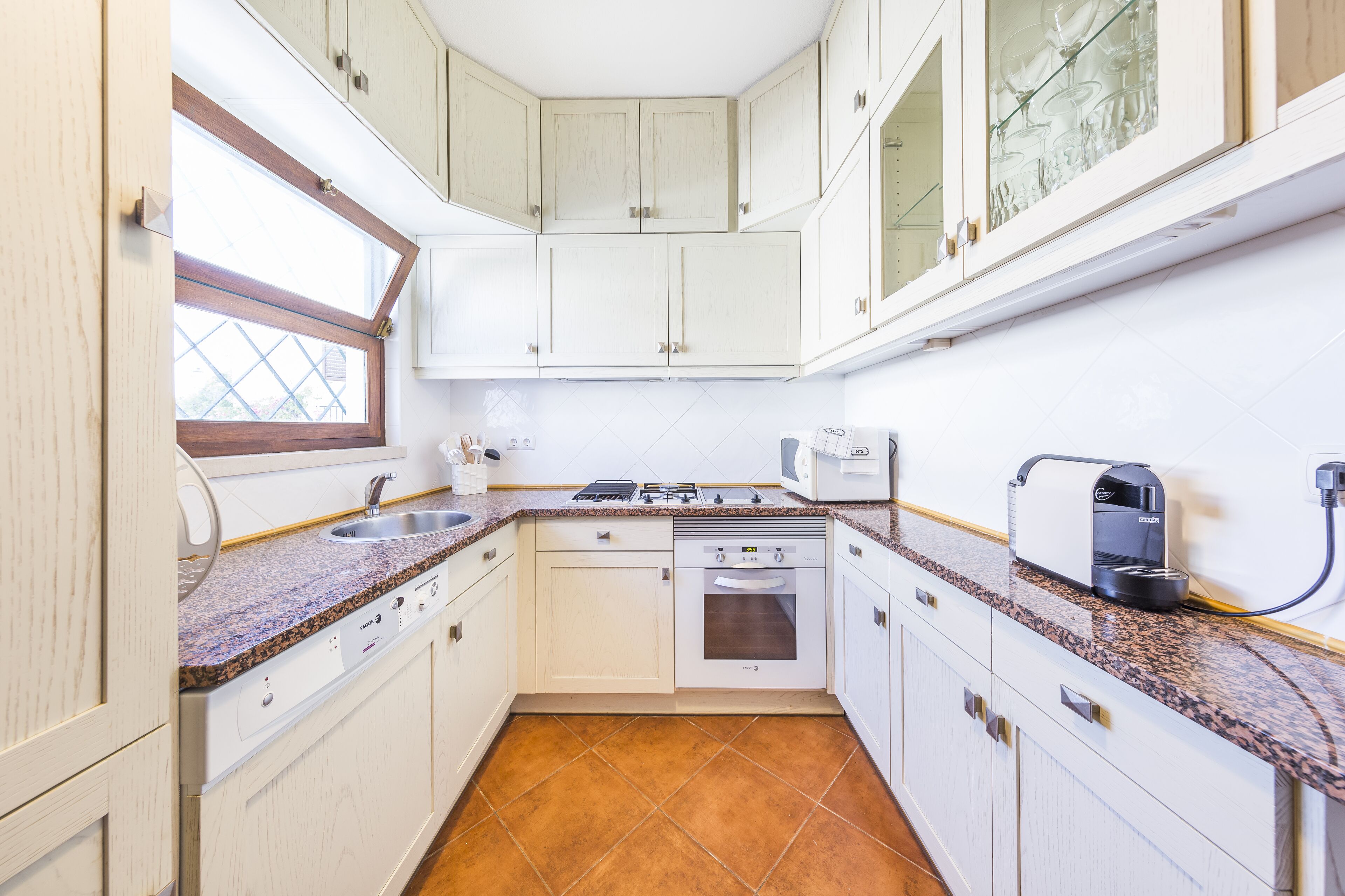 A compact kitchen with white cabinets, a built-in oven, a coffee maker, and a window.