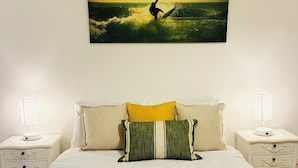 Ein ordentlich gemachtes Bett mit zwei Kissen, einem gefalteten Handtuch und einem Gemälde eines Surfers auf einer Welle darüber.